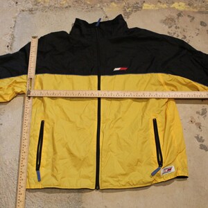 Pu&ograve; includere: Una giacca a vento gialla e nera con chiusura a zip. La giacca ha un piccolo logo sul petto sinistro e due tasche con zip sui lati.