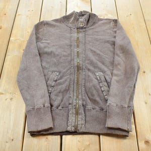 May include: A faded brown zip-up jacket with a ribbed collar, cuffs, and hem. The jacket features a full-length brass zipper and two front pockets. The fabric has a textured appearance.