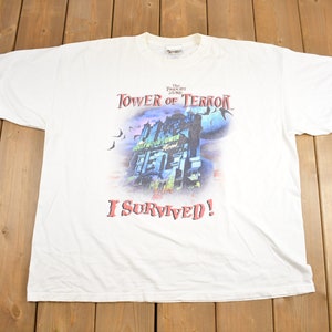 May include: White t-shirt with a graphic of the Tower of Terror ride at Disney World. The graphic shows the haunted hotel with bats flying around it. The text "The Twilight Zone Tower of Terror 2004" and "I Survived!" are printed on the shirt.