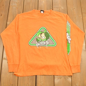 May include: Orange long-sleeve shirt with a green triangle graphic featuring a cartoon character skateboarding. The text "Tech Deck" is printed on the right sleeve.