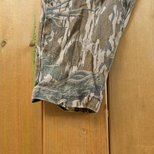 May include: Close-up of a section of camouflage pants. The fabric features a pattern of brown, green, and gray tones, creating a woodland effect. The pants are made of a durable material, suitable for outdoor activities.