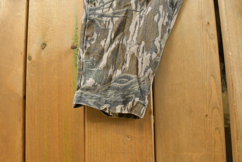May include: Close-up of a section of camouflage pants. The fabric features a pattern of brown, green, and gray tones, creating a woodland effect. The pants are made of a durable material, suitable for outdoor activities.