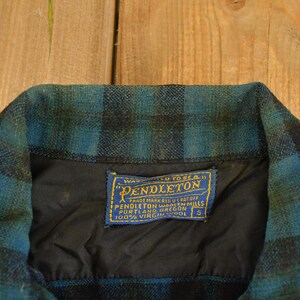 Op de afbeelding: Detail van de kraag van een vintage Pendleton wollen overhemd. De kraag heeft een blauw en zwart ruitpatroon. Een blauw en goud label met het Pendleton logo en tekst is in de zwarte voering genaaid. Het overhemd is maat S.