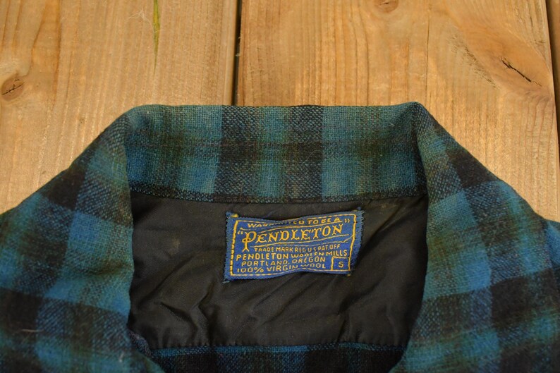 Op de afbeelding: Detail van de kraag van een vintage Pendleton wollen overhemd. De kraag heeft een blauw en zwart ruitpatroon. Een blauw en goud label met het Pendleton logo en tekst is in de zwarte voering genaaid. Het overhemd is maat S.