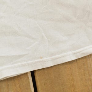 May include: Close-up of a white fabric, possibly a t-shirt, with visible texture and slight wrinkles. The edge of the fabric is neatly hemmed with a visible seam. The fabric rests on a wooden surface.