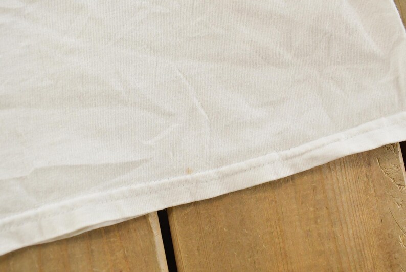 May include: Close-up of a white fabric, possibly a t-shirt, with visible texture and slight wrinkles. The edge of the fabric is neatly hemmed with a visible seam. The fabric rests on a wooden surface.