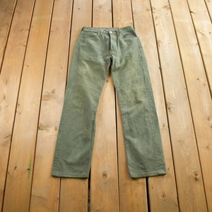 Op de afbeelding: Olijfgroene denim jeans met een knoopsluiting, klassiek vijf-zakken ontwerp en een rechte pijp. De jeans liggen plat op een houten oppervlak en tonen hun versleten uiterlijk en vintage stijl.