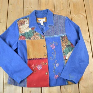 May include: A patchwork jacket with a blue denim body and floral, tan, and red panels. The jacket features embroidered floral accents and gold-colored buttons. The collar and sleeves are solid blue.