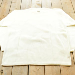 May include: Cream-colored long-sleeved shirt with a crew neck. The shirt has a small logo on the upper back and vertical seams down the front and back. The fabric appears to be a soft, textured material.