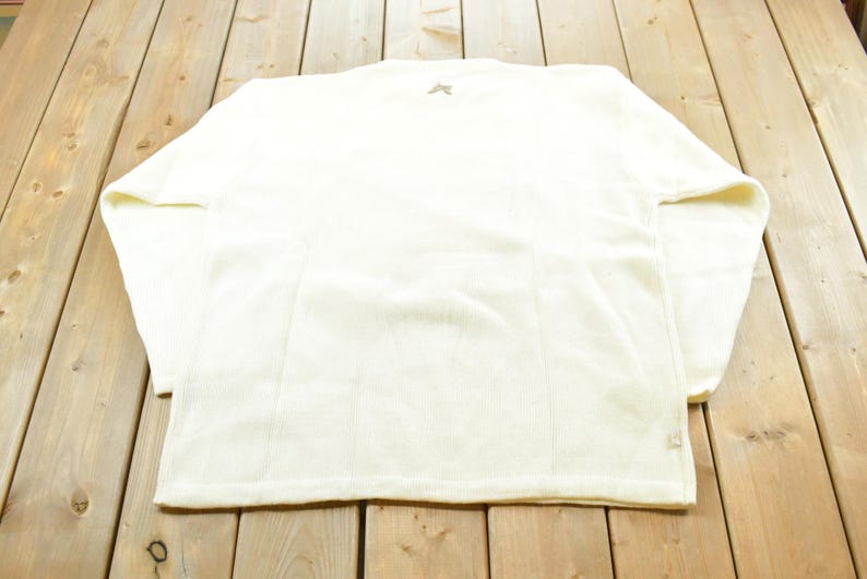 May include: Cream-colored long-sleeved shirt with a crew neck. The shirt has a small logo on the upper back and vertical seams down the front and back. The fabric appears to be a soft, textured material.