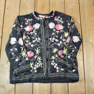 Puede incluir: Una camisa negra de manga larga con botones y bordados florales. El bordado presenta flores rosas, blancas y amarillas.
