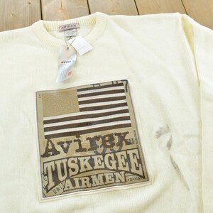 May include: Cream-colored crew neck sweatshirt with a rectangular patch featuring an American flag design and the text "Avirex Tuskegee Airmen". The sweatshirt has a ribbed texture and a tag attached to the collar.