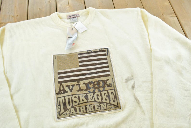 May include: Cream-colored crew neck sweatshirt with a rectangular patch featuring an American flag design and the text "Avirex Tuskegee Airmen". The sweatshirt has a ribbed texture and a tag attached to the collar.