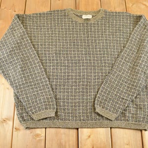 May include: A long-sleeve crew neck jumper featuring a textured, repeating geometric pattern in shades of olive green, charcoal grey, and cream. The garment has a ribbed knit collar, cuffs, and hem, with a small white brand label at the neckline.