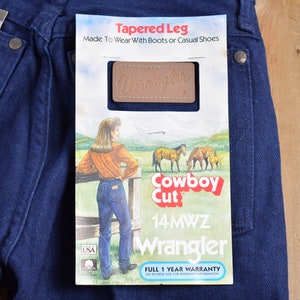 May include: A pair of dark blue denim jeans with a Wrangler label and a tag that reads "Tapered Leg, Made To Wear With Boots or Casual Shoes, Cowboy Cut, 14MWZ, Wrangler, Full 1 Year Warranty".
