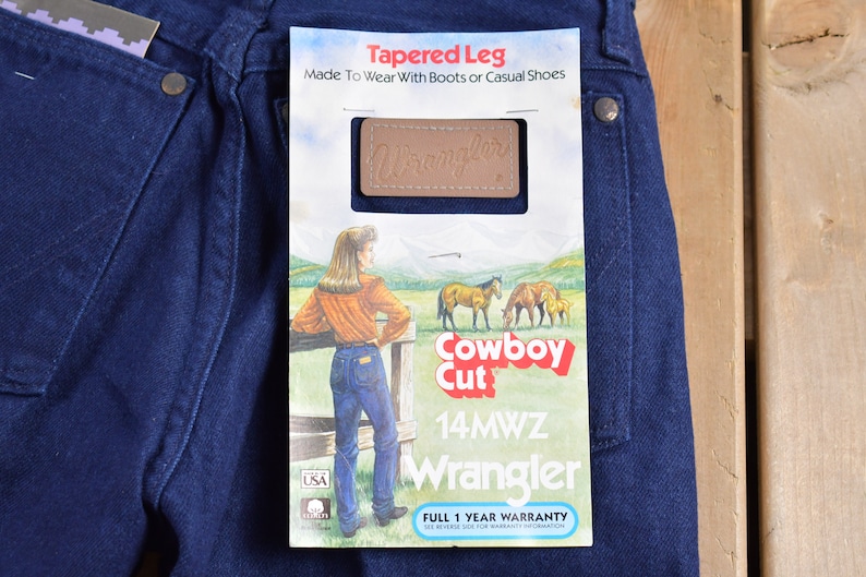 May include: A pair of dark blue denim jeans with a Wrangler label and a tag that reads "Tapered Leg, Made To Wear With Boots or Casual Shoes, Cowboy Cut, 14MWZ, Wrangler, Full 1 Year Warranty".
