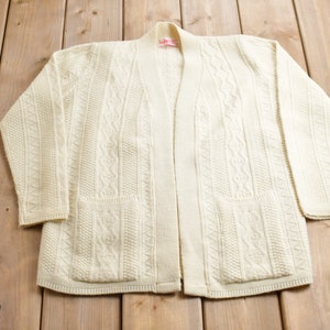 Puede incluir: Un cárdigan blanco de punto de cable con manga larga y dos bolsillos. El cárdigan tiene un diseño sencillo y está hecho de un material suave y cómodo.