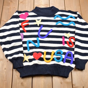 May include: A black and white striped sweater with the words "FUN IS USA" spelled out in colorful yarn. The letters are decorated with hearts and stars.