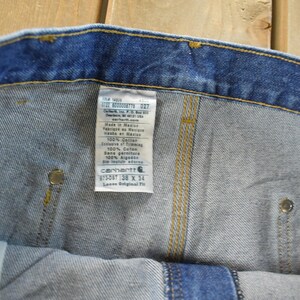 K&ouml;nnte beinhalten: Nahaufnahme einer blauen Jeans mit Etikett. Das Etikett enth&auml;lt den Markennamen Carhartt, die Gr&ouml;&szlig;e 38 x 34 und weitere Details. Die Jeans hat gelbe N&auml;hte und Metallnieten.