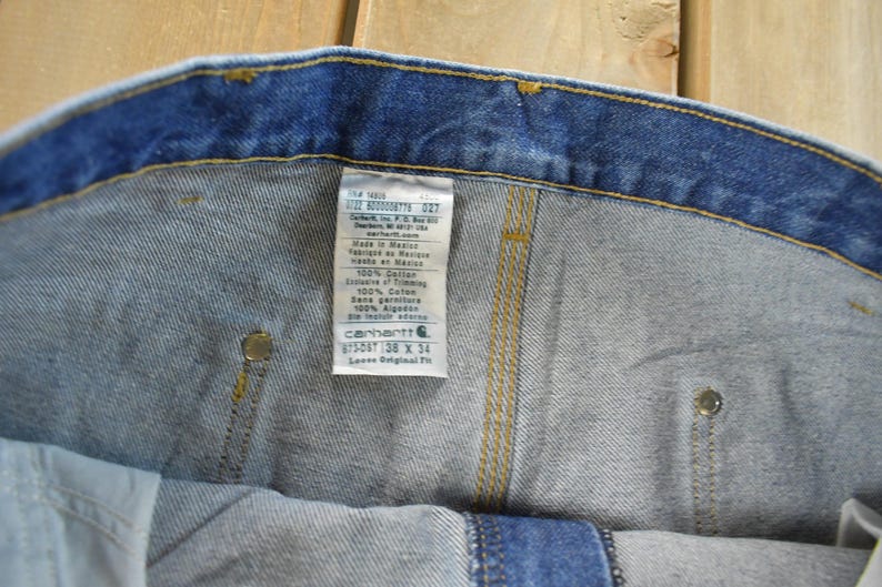 K&ouml;nnte beinhalten: Nahaufnahme einer blauen Jeans mit Etikett. Das Etikett enth&auml;lt den Markennamen Carhartt, die Gr&ouml;&szlig;e 38 x 34 und weitere Details. Die Jeans hat gelbe N&auml;hte und Metallnieten.