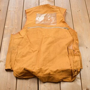 May include: A brown canvas vest with a zippered pocket and a clear plastic pocket on the back. The vest has two side pockets with button closures.