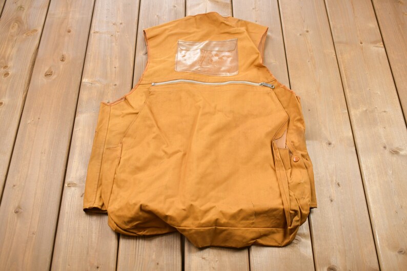 May include: A brown canvas vest with a zippered pocket and a clear plastic pocket on the back. The vest has two side pockets with button closures.