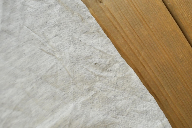 Op de afbeelding: Een lichtgrijze gem&ecirc;leerde stof met een zachte, gespikkelde textuur en zichtbare vouwen. Het materiaal oogt lichtgewicht en enigszins gekreukt, met een klein, donker vlekje nabij het midden van het zichtbare oppervlak.