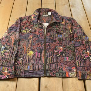 May include: A long-sleeve jacket with a full-length front zipper, a collar, and two chest pockets. The fabric features a dark background with a colorful, intricate pattern of giraffes, stylized figures, and geometric shapes in yellow, pink, and blue.