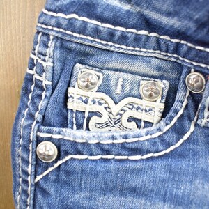 K&ouml;nnte beinhalten: Nahaufnahme einer blauen Jeanshose mit wei&szlig;en N&auml;hten. Die Jeans hat eine Tasche mit einem wei&szlig;en, dekorativen Design und silbernen Kn&ouml;pfen.
