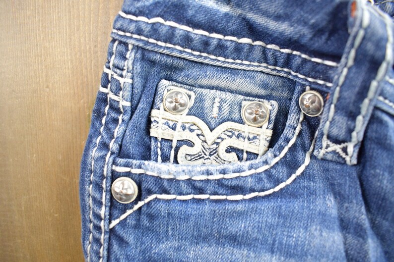 K&ouml;nnte beinhalten: Nahaufnahme einer blauen Jeanshose mit wei&szlig;en N&auml;hten. Die Jeans hat eine Tasche mit einem wei&szlig;en, dekorativen Design und silbernen Kn&ouml;pfen.