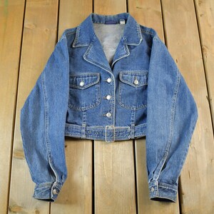 May include: A vintage blue denim jacket with a collared neckline, button-down front, and two chest pockets. The jacket features long sleeves with buttoned cuffs and a belt at the waist. The jacket is approximately 20 inches long.