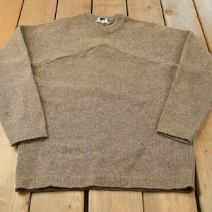 Könnte beinhalten: Ein langärmeliger Pullover mit Rundhalsausschnitt in einem einfarbigen Beigeton. Das Kleidungsstück weist eine markante horizontale Naht im Brustbereich auf, die eine geschwungene, bogenförmige Linie bildet. Das Strickmuster ist einheitlich.
