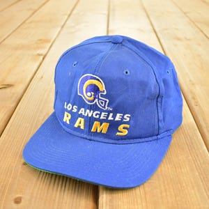 May include: A blue baseball cap with the Los Angeles Rams logo. The cap features a yellow and white helmet graphic and the team name in yellow lettering. The brim is blue with a green underside.