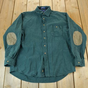 Peut inclure: Une chemise boutonnée vert teal à manches longues et col. La chemise présente des coudières beiges et une poche poitrine. La chemise est posée à plat sur une surface en bois.