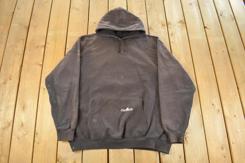 May include: A dark gray hooded sweatshirt with a front pocket and drawstring. The Carhartt logo is visible on the front pocket. The sweatshirt is made of a thick, durable material.
