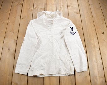 May include: A white long-sleeved shirt with a hood and a blue eagle patch on the right sleeve. The shirt has a button-up closure at the neck.