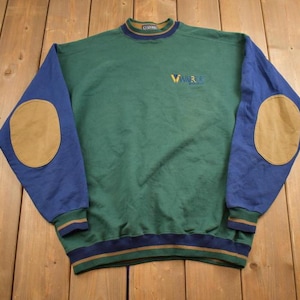 Puede incluir: Una sudadera verde y azul con parches marrones en los codos. La sudadera tiene un cuello y puños de canalé. El texto "VIMAR" está bordado en la parte delantera de la sudadera.
