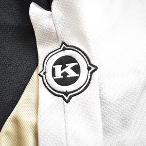 Puede incluir: Jersey negro y blanco con una franja dorada y un logotipo negro y blanco con la letra "K" en el centro.