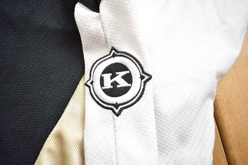 Puede incluir: Jersey negro y blanco con una franja dorada y un logotipo negro y blanco con la letra "K" en el centro.