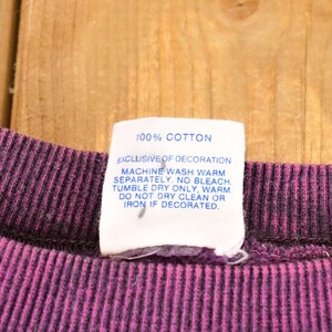May include: A dark purple ribbed crew neck collar with a white fabric care tag attached. The tag reads 100% COTTON, EXCLUSIVE OF DECORATION, MACHINE WASH WARM SEPARATELY. NO BLEACH. TUMBLE DRY ONLY, WARM. DO NOT DRY CLEAN OR IRON IF DECORATED.