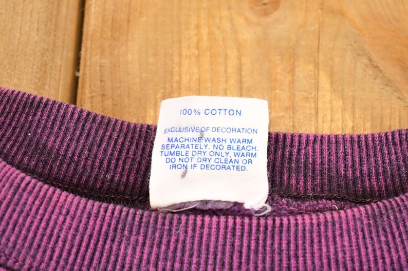 May include: A dark purple ribbed crew neck collar with a white fabric care tag attached. The tag reads 100% COTTON, EXCLUSIVE OF DECORATION, MACHINE WASH WARM SEPARATELY. NO BLEACH. TUMBLE DRY ONLY, WARM. DO NOT DRY CLEAN OR IRON IF DECORATED.