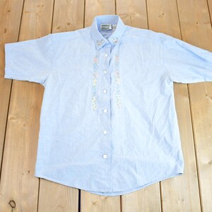 Puede incluir: Una camisa de manga corta de color azul claro con detalles florales bordados.