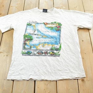 Puede incluir: Camiseta blanca con un mapa del Mar Caribe, con las inscripciones "St. Maarten" y "Antigua". El diseño presenta una combinación de colores azul y verde con ilustraciones de elementos tropicales. Cuello redondo y mangas cortas.