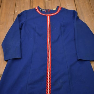 Puede incluir: Un vestido azul con ribete rojo y blanco alrededor del escote y en la parte delantera. El vestido tiene mangas largas.