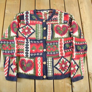 May include: A colorful cardigan sweater with a patchwork design. The sweater features a variety of patterns including hearts, flowers, and geometric shapes in red, green, white, and navy blue. It has a button-up front and a navy blue trim.