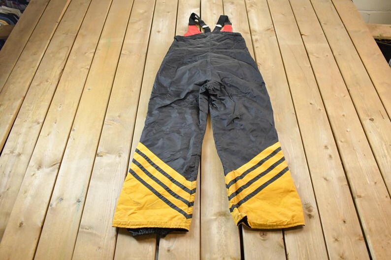 May include: Black ski overalls with yellow and black stripes on the legs. The straps are red and black.