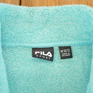 May include: Close-up of a turquoise fleece jacket collar. The black label reads "FILA SPORT" in white text. A smaller tag indicates size M 10/12 and "MADE IN VIETNAM". The soft, textured fabric is visible.