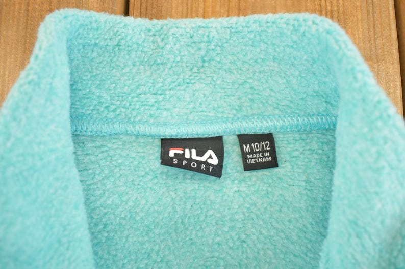 May include: Close-up of a turquoise fleece jacket collar. The black label reads "FILA SPORT" in white text. A smaller tag indicates size M 10/12 and "MADE IN VIETNAM". The soft, textured fabric is visible.
