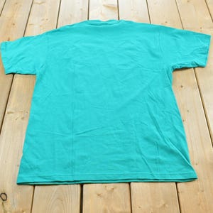 May include: A solid teal short-sleeved t-shirt with a crew neckline. The garment is laid flat and features a plain, unpatterned design on the back, showcasing the consistent vibrant color and standard construction of the cotton fabric throughout.
