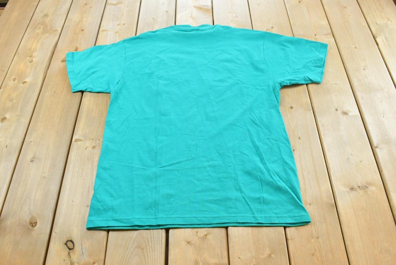 May include: A solid teal short-sleeved t-shirt with a crew neckline. The garment is laid flat and features a plain, unpatterned design on the back, showcasing the consistent vibrant color and standard construction of the cotton fabric throughout.